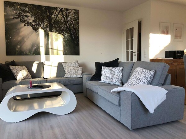 Una sala de estar moderna con sofás grises, cojines estampados, una mesa de centro blanca y un cuadro abstracto grande en la pared.