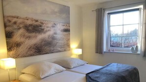 A double bed with white linens, a bedside table with a lamp, a large abstract painting on the wall, and a window with curtains.