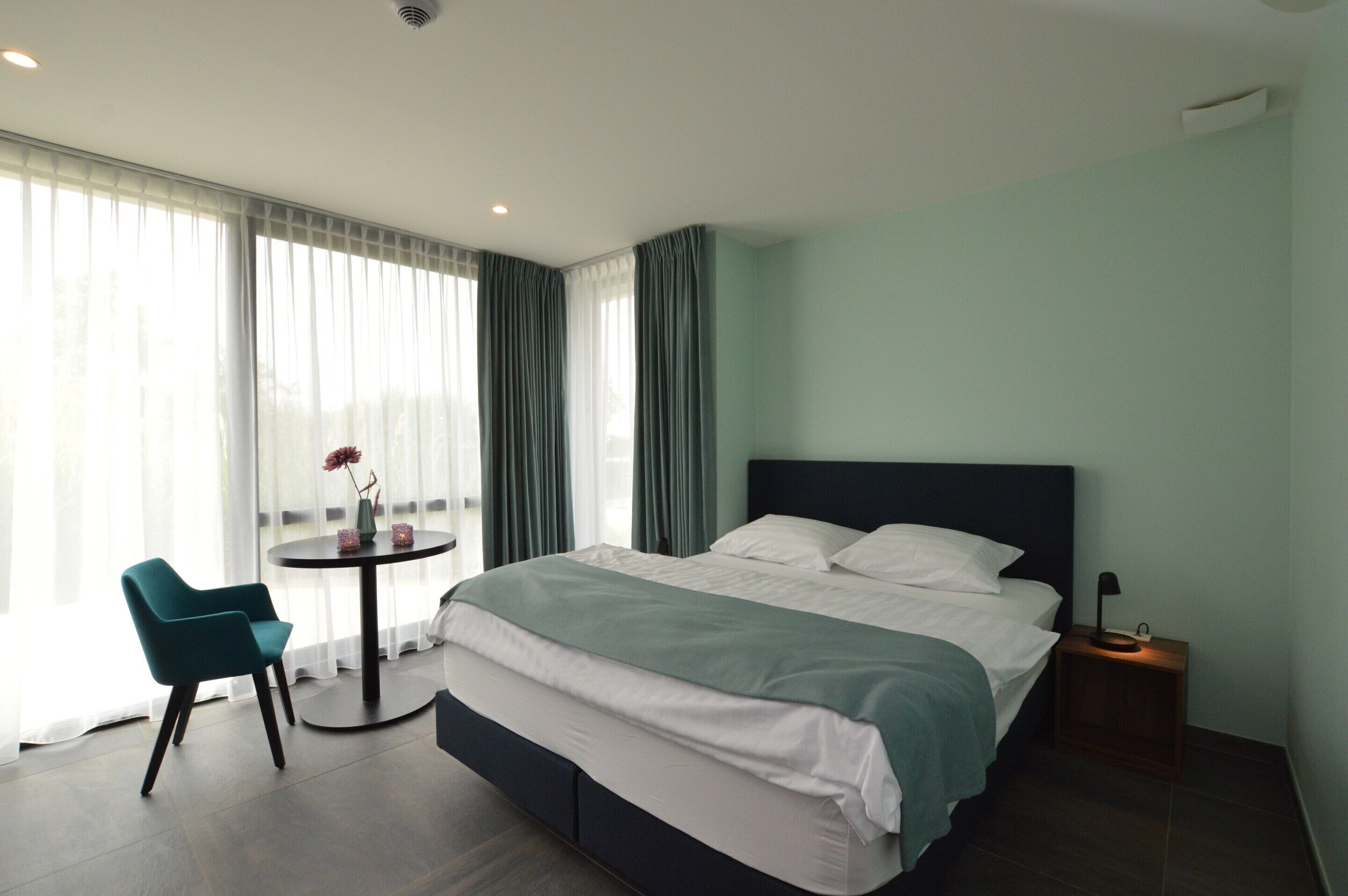 A modern hotel room with a large bed, a round table with a chair, and a view of the outdoors through sheer curtains.