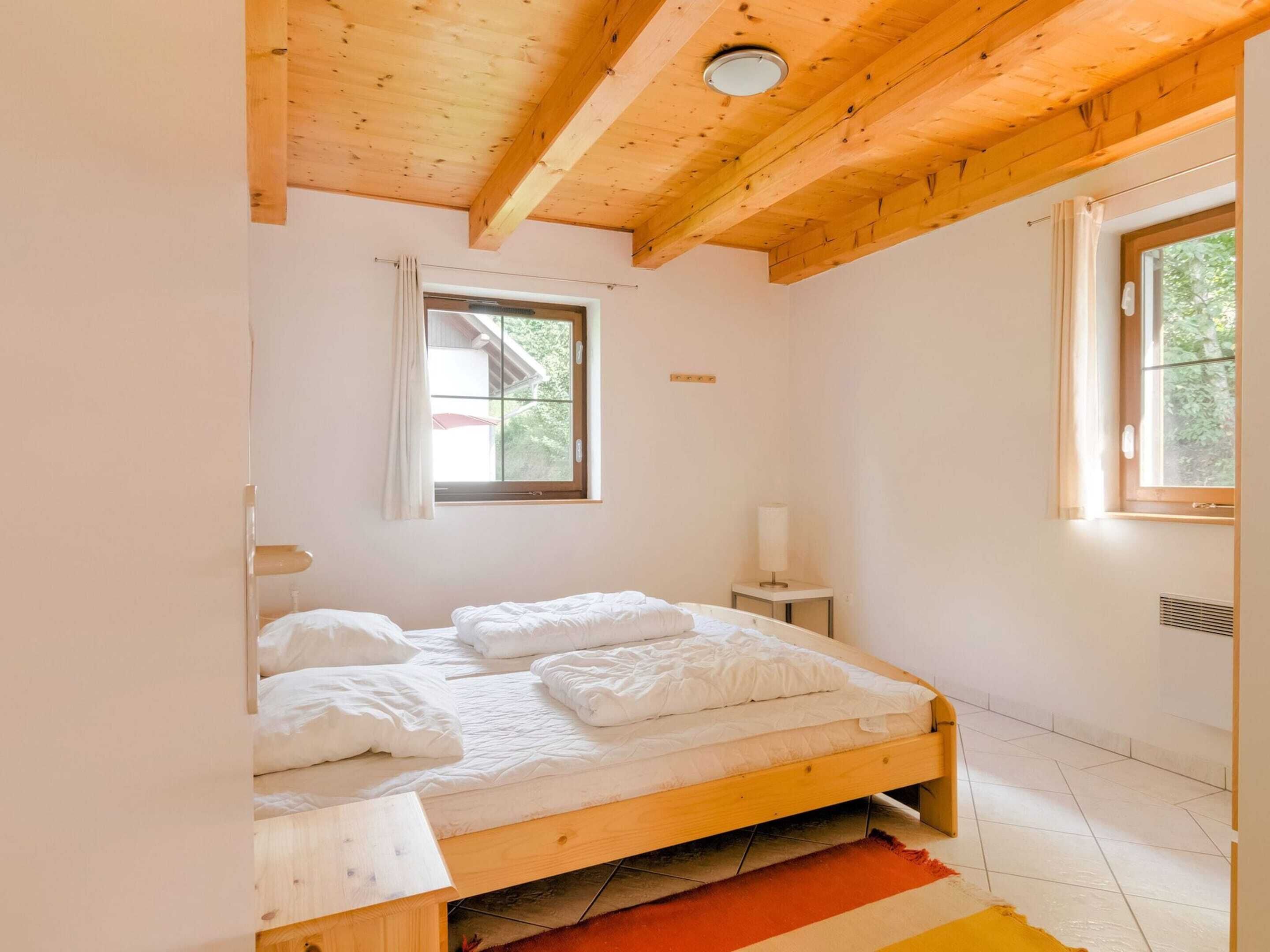 A single bed with white bedding in a room with wooden beams on the ceiling, a window with curtains, and a small bedside table with a lamp.