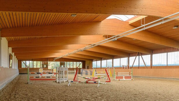 Arena equestre coberta com vigas de madeira, chão de areia e diversos obstáculos de salto.