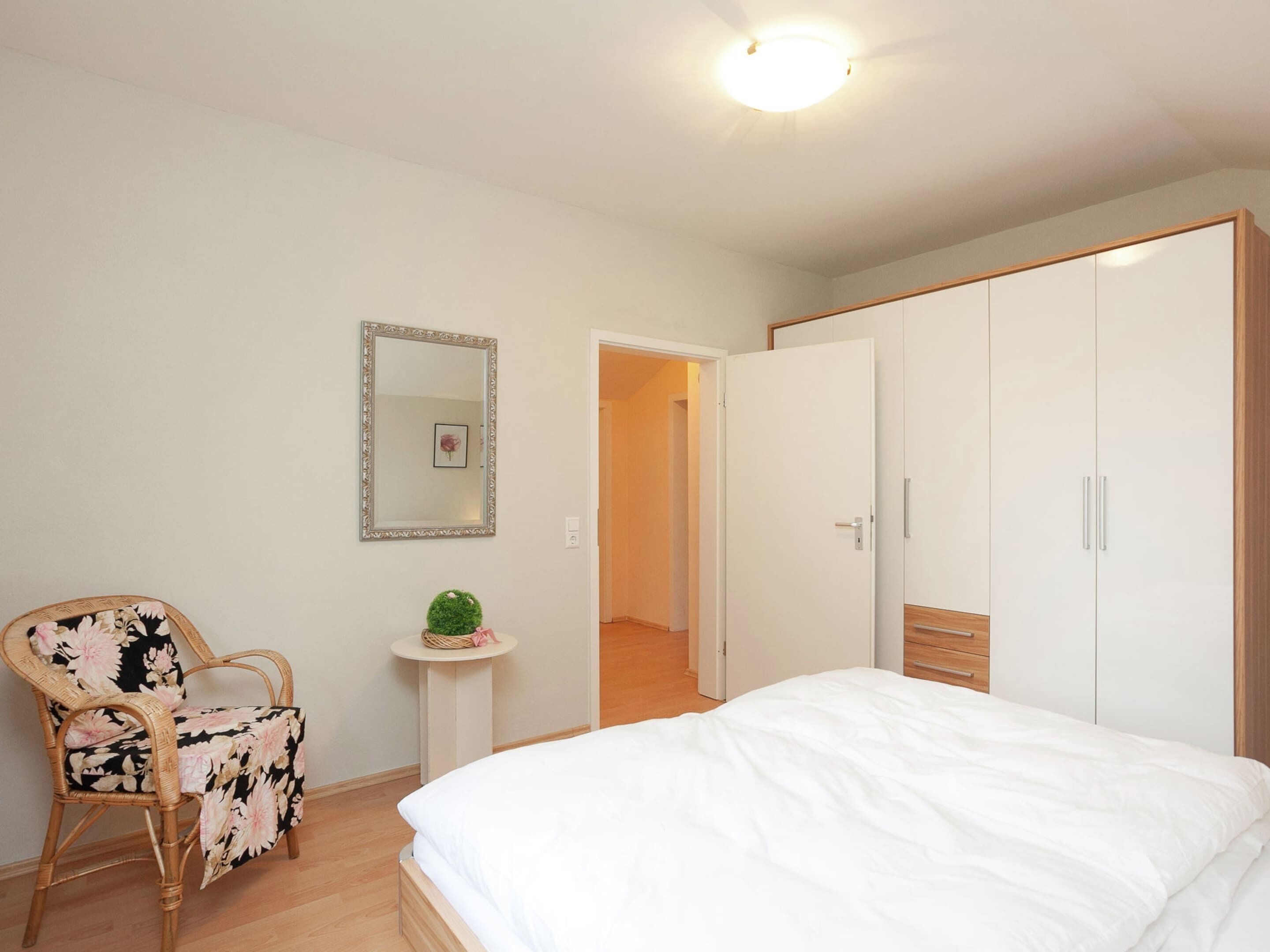 A bedroom with a bed, a chair, a small table with a plant, a mirror, and a wardrobe.