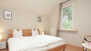 A neatly made bed in a room with a wooden headboard, a small bedside table with a lamp, a window with a view of trees, and a framed picture on the wall. - Ferienhaus in Rechenberg-bienemuehle (Rechenberg-Bienenmuehle)