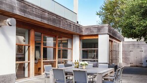 Un patio al aire libre con una mesa grande y sillas, rodeado de amplios ventanales y una terraza de madera.