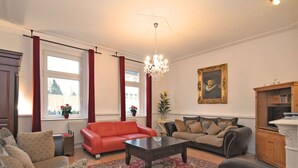A living room with a red sofa, a patterned armchair, a dark coffee table, and a beige sofa with patterned cushions. There is a chandelier hanging from the ceiling, a framed portrait on the wall, and a wooden cabinet with a decorative top. - Heritage Villa With Home Cinema, Sauna (Treuen)
