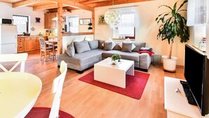 A spacious living room with a large sofa, a coffee table, and a TV. There is a kitchen area with wooden cabinets and a microwave. A dining table with chairs is also visible. - Ferienhaus in Thorstorf mit Garten (Warnow)