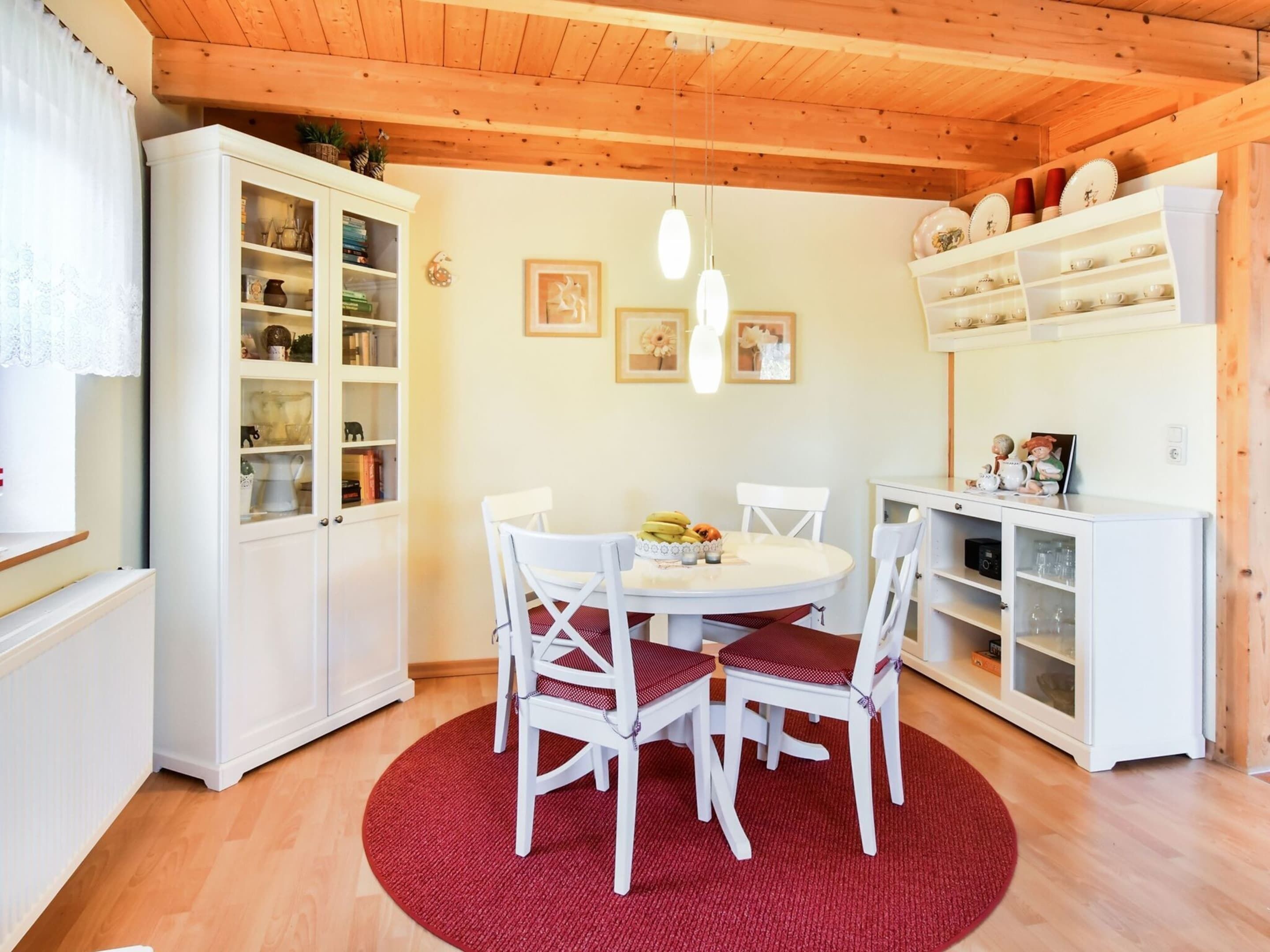 A cozy dining area with a round table, white chairs, and a red rug. There are cabinets with glass doors on the left and a shelf with dishes on the right.