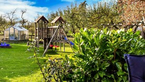 A wooden play structure with a slide and climbing elements, a greenhouse, and a well-maintained lawn. - Ferienhaus in Thorstorf mit Garten (Warnow)