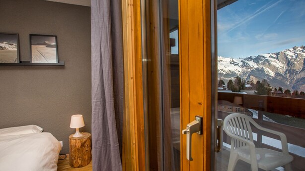 Une chambre d’hôtel avec un lit, une table de chevet, une lampe, un tabouret en bois, un balcon avec une chaise et une vue sur la montagne.