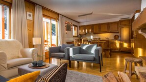 Un salon chaleureux avec un canapé, un fauteuil et un coin repas agrémenté de meubles en bois, et une cuisine en arrière-plan.