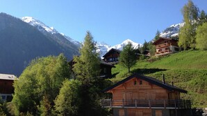 Un chalet en bois doté d’un balcon, entouré d’une végétation luxuriante et avec des montagnes enneigées en toile de fond.