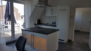 Ett modernt kök med en köksö i mitten, en mikrovågsugn och en inbyggd ugn. Det finns en skjutdörr i glas som leder ut till ett utomhusområde med en stol och ett bord.