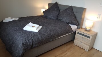 Una cama bien hecha con una colcha oscura estampada y almohadas, una toalla blanca y una mesita de noche de madera con una lámpara.