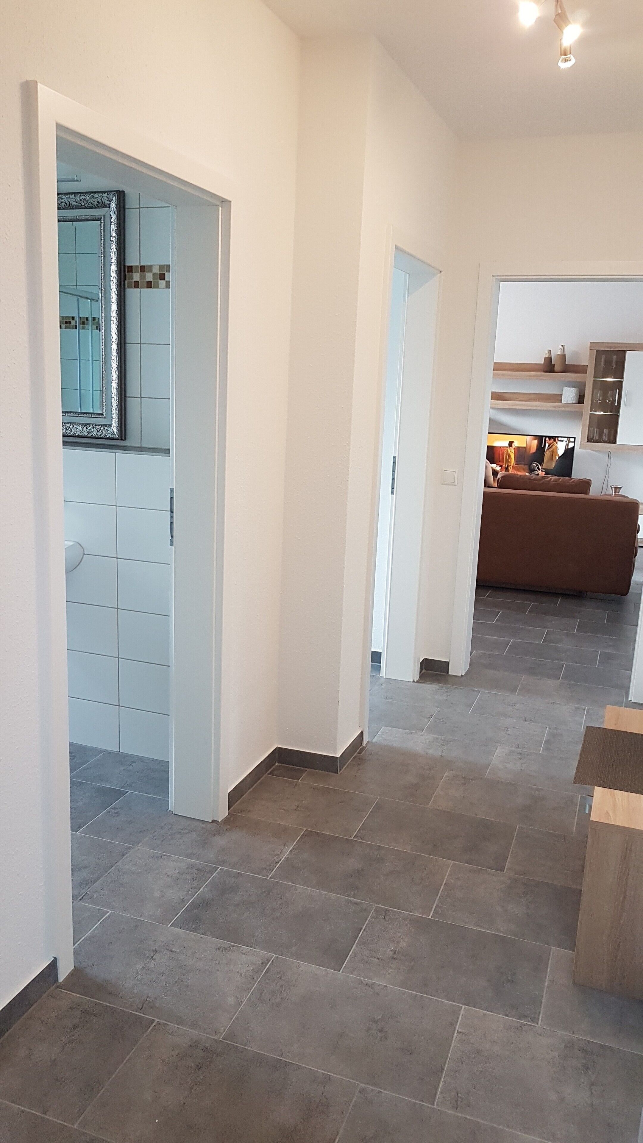 Ein geräumiger Korridor mit Fliesenboden, der in einen Wohnbereich mit braunem Sofa und Fernseher führt.