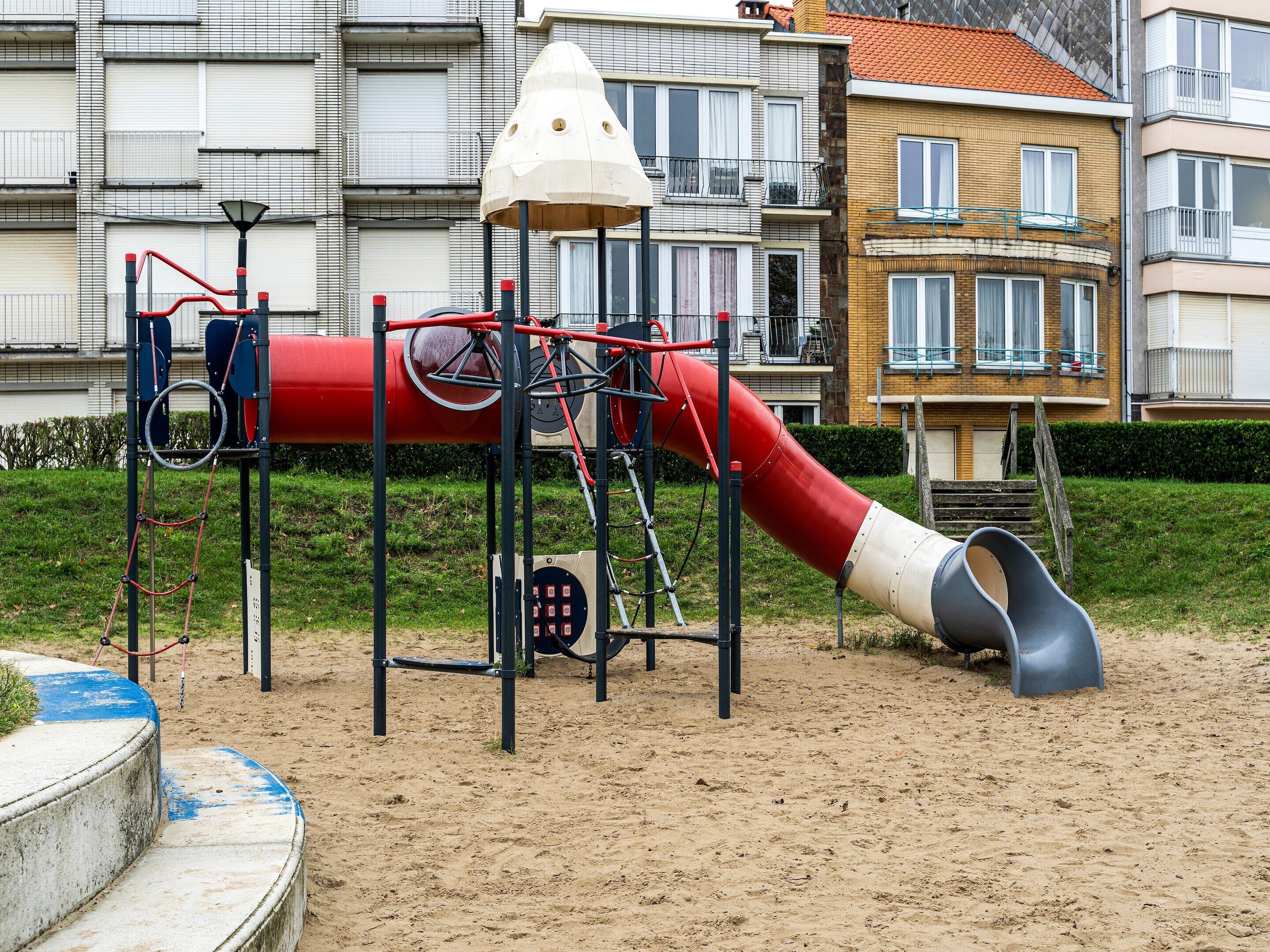 Un parque infantil con un tobogán rojo y estructuras para escalar, con edificios residenciales de fondo.