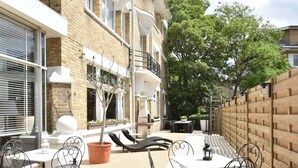 An outdoor patio with a table and chairs, a potted tree, and a brick building in the background. - Former Hotel in the Centre and Near the Beach in Panne (De Panne)