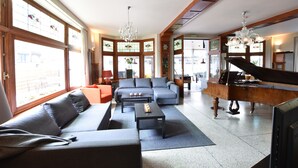 A spacious living room with a grand piano, a sectional sofa, and a large window with stained glass. - Former Hotel in the Centre and Near the Beach in Panne (De Panne)