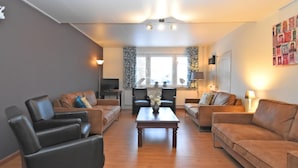 A living room with two sofas, a coffee table, and a TV. There is a window with curtains and a radiator.