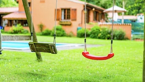 A wooden swing set in a grassy area with a pool and a building in the background. - Cosy and Snug Holiday Home With Joint Swimming Pool (Tessenderlo)