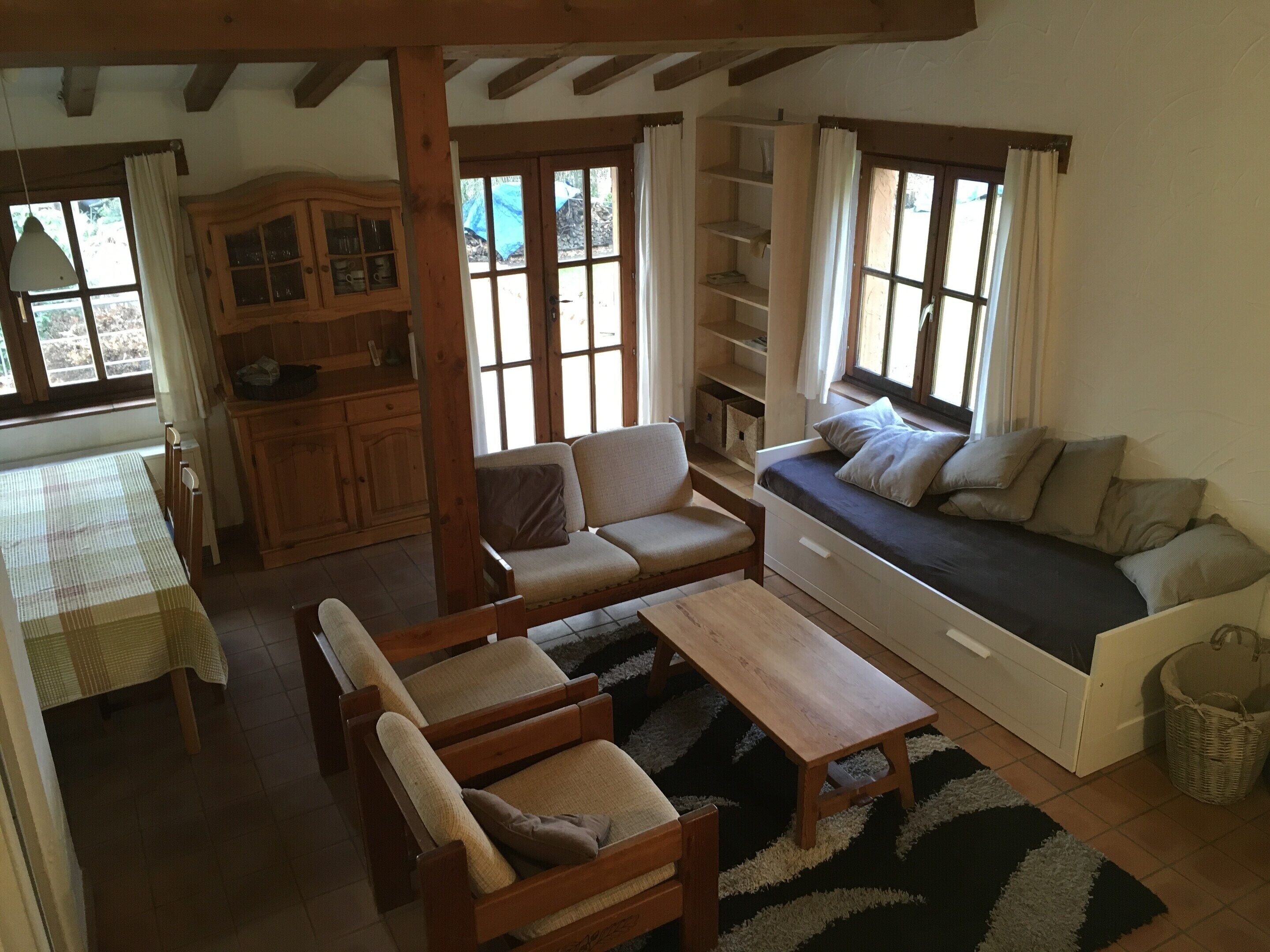 A cozy living room with a sofa, chairs, a table, and a built-in bench with storage. There are large windows with curtains, a wooden cabinet, and a small kitchen area.