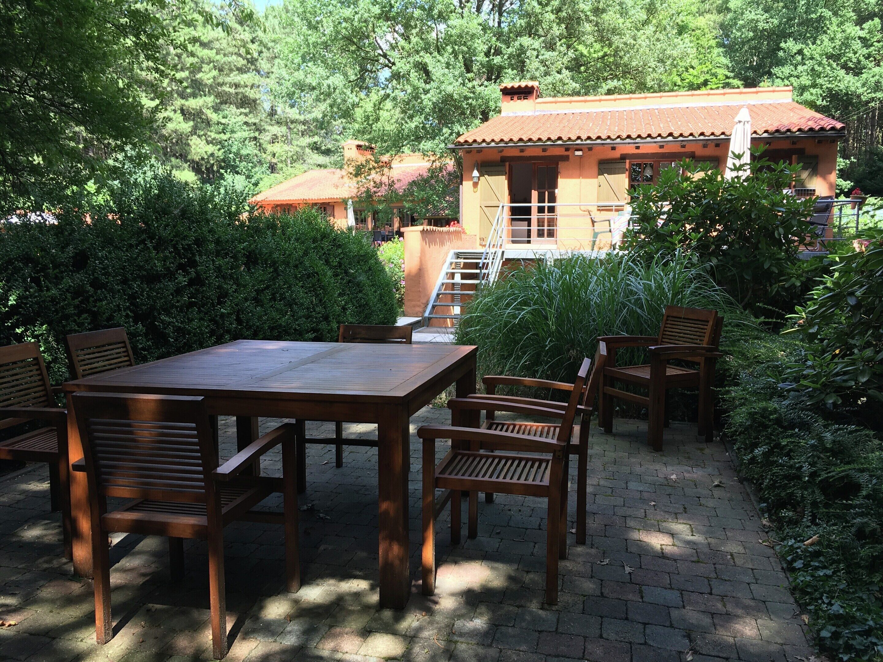 Une terrasse extérieure avec une table et des chaises en bois, entourée d’une végétation luxuriante et, au fond, un bâtiment doté d’un toit en tuiles.