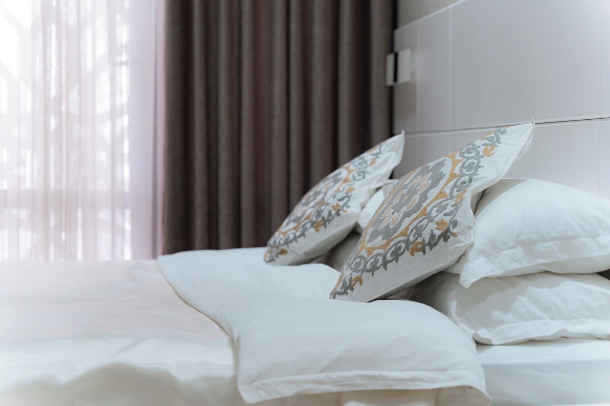 A neatly made bed with white bedding and patterned pillows, a window with curtains, and a white wall.