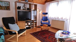 Un salon avec un plancher en bois, un téléviseur, un fauteuil bleu, une table basse en verre et un tapis à motifs.