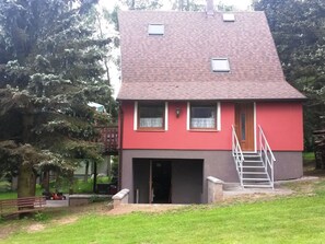 A two-story house with a red and grey exterior, a front door, and a small front yard with a bench and a ladder. - House in Bohemian Near Ski Slope (Blatno)