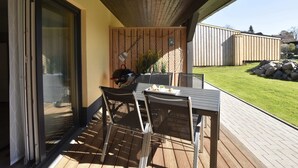 An outdoor patio with a table and chairs, a grill, and a view of a backyard with a wooden fence and a lawn.