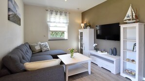 A modern living room with a grey sofa, a white coffee table, a TV mounted on the wall, and a bookshelf.