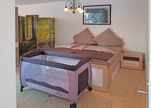 Une chambre à coucher avec un lit, une petite table de chevet, une commode, un parc pour bébé, une lampe fixée au mur et un tableau encadré.