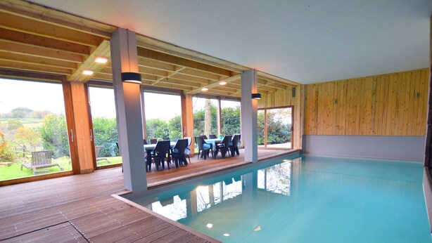 An indoor swimming pool with wooden decking, a covered seating area with chairs, and large windows offering a view of the outdoors. - Rustic Farmhouse With Pool (Waimes)