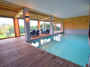 An indoor swimming pool with wooden decking, a covered seating area with chairs, and large windows offering a view of the outdoors.