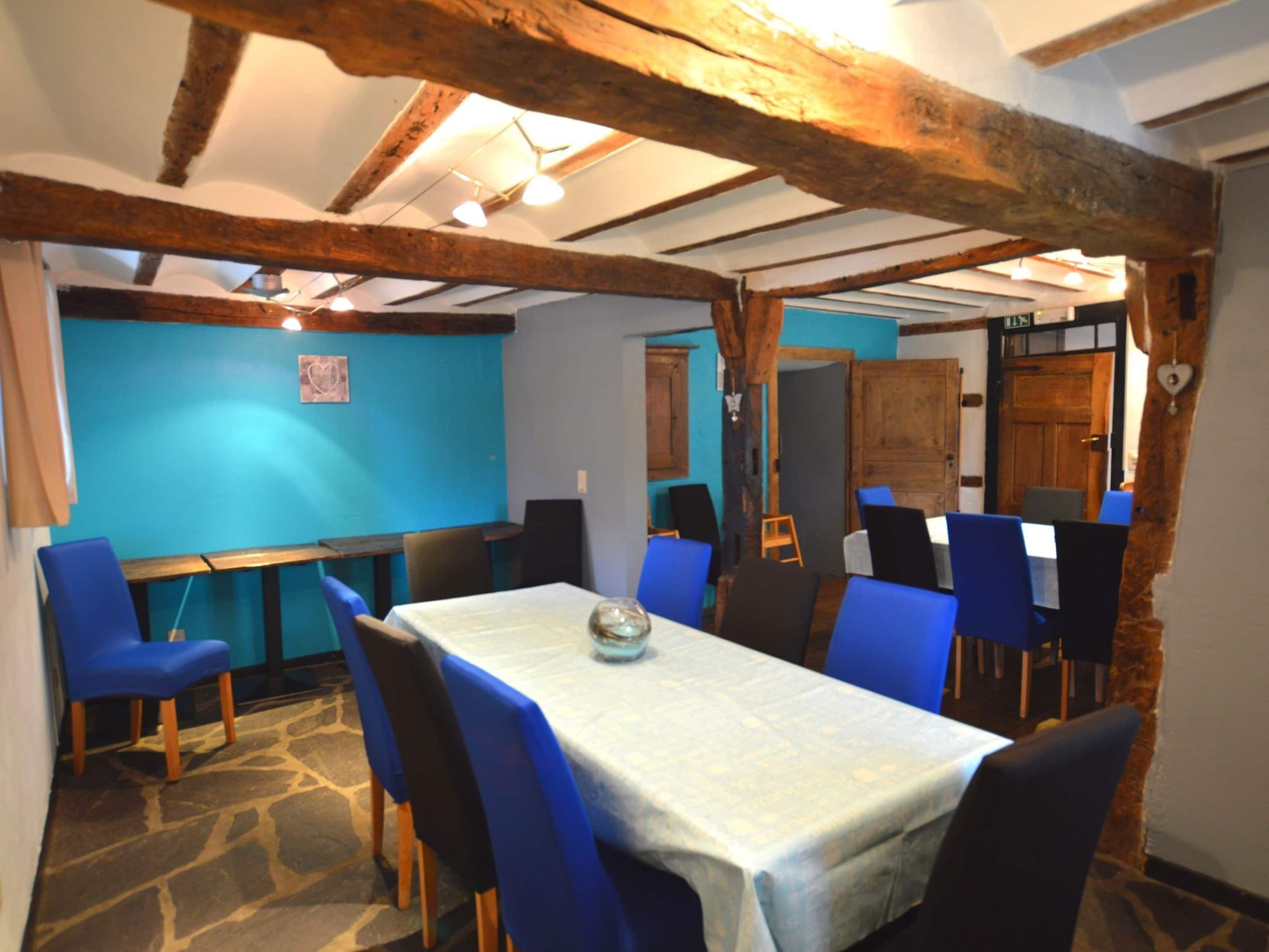 A dining area with a table set for four, surrounded by blue chairs, under exposed wooden beams.