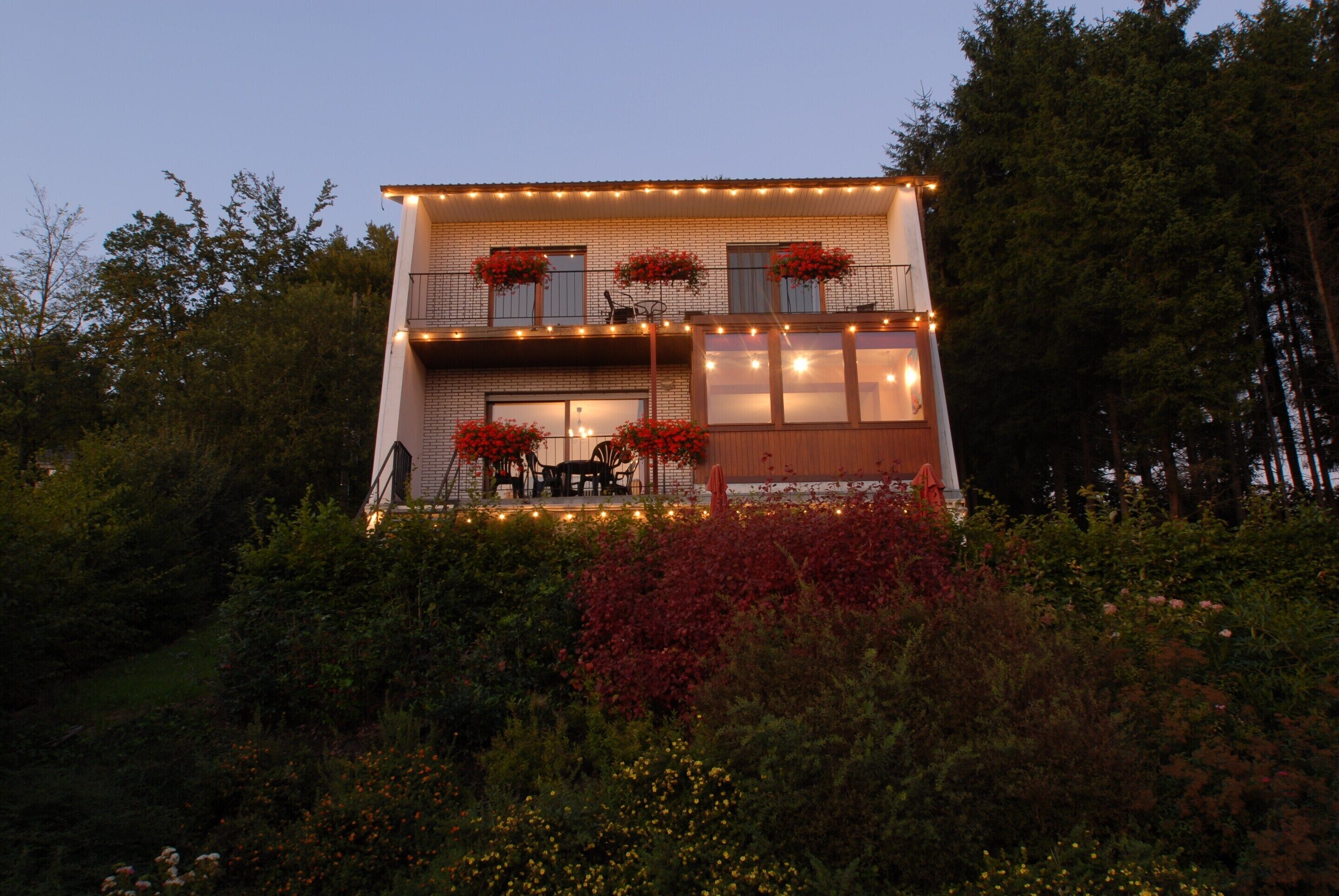 A two-story building with a balcony, surrounded by dense greenery and flowering plants.