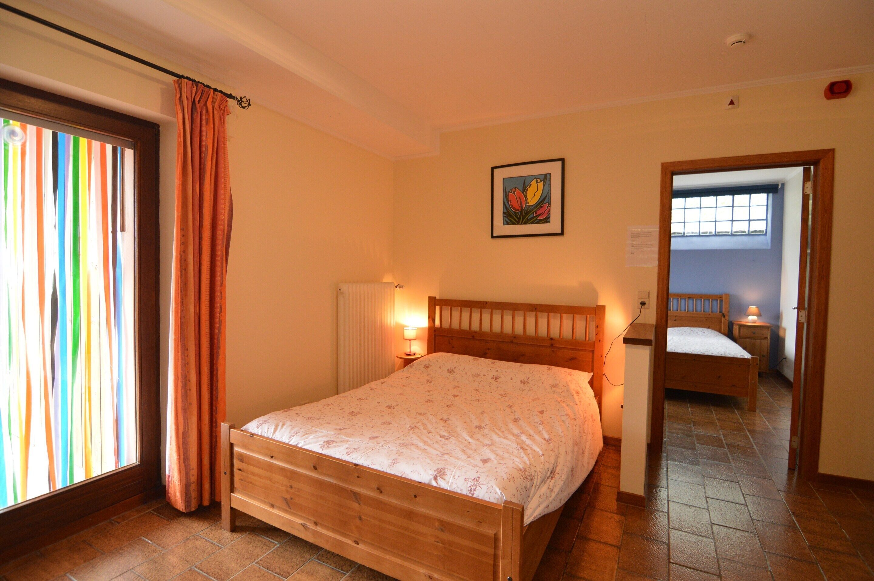 Un dormitorio con cama de madera, una ventana con cortinas, una lámpara de noche y un cuadro en la pared.