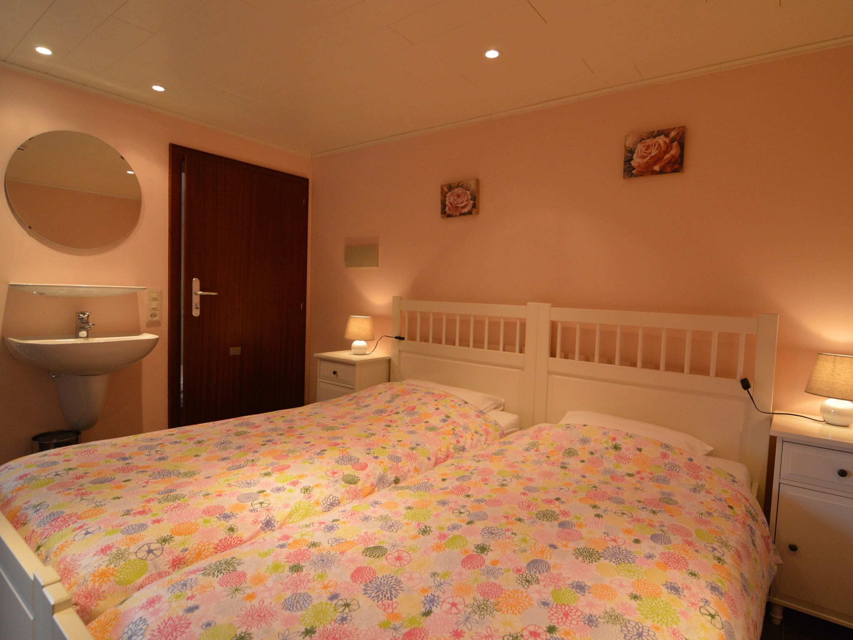A bedroom with a bed, a bedside table, a nightstand, a mirror, and a wall-mounted sink.