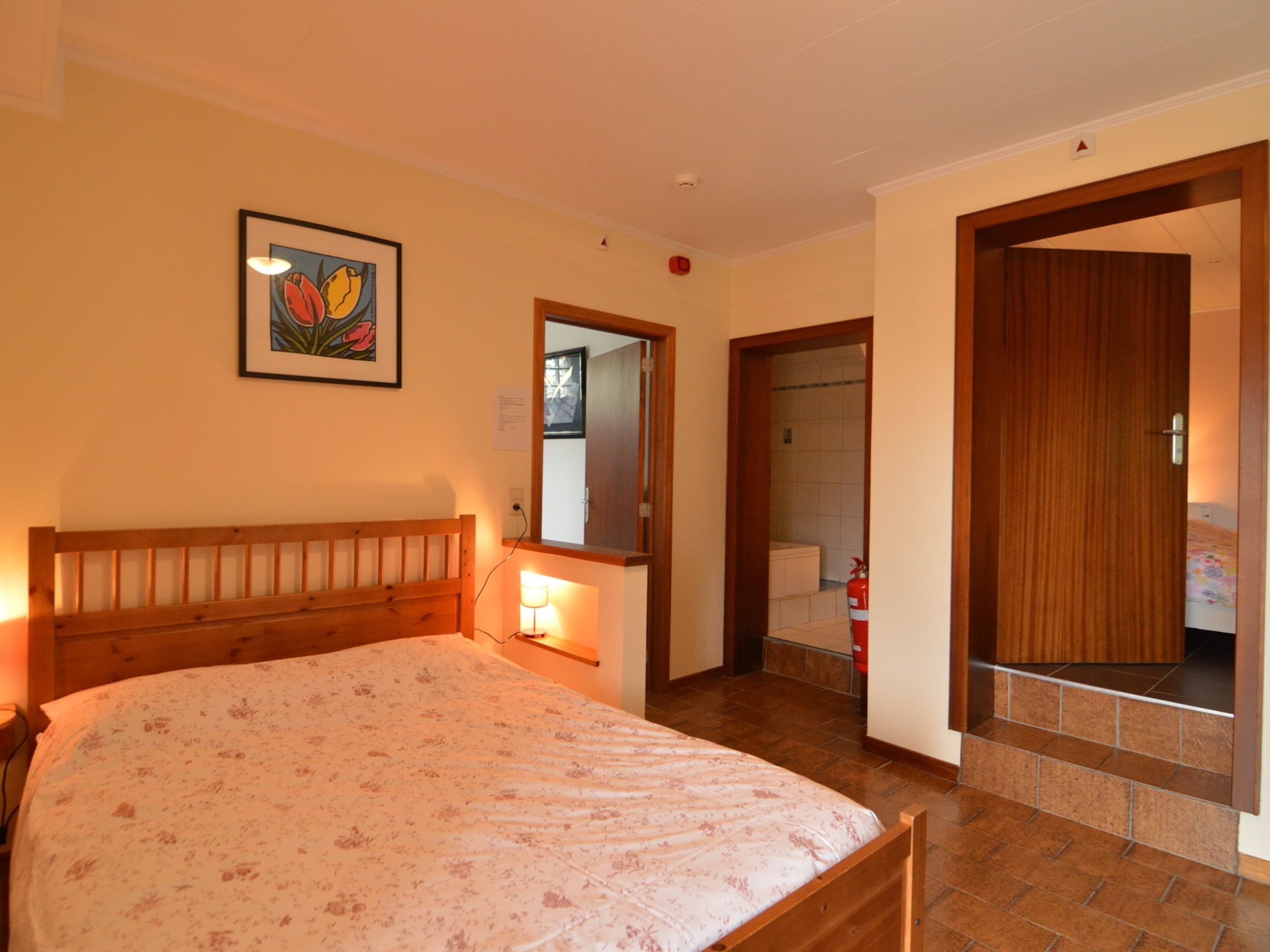 A bedroom with a wooden bed, a bedside lamp, a framed picture on the wall, and a door leading to a bathroom.