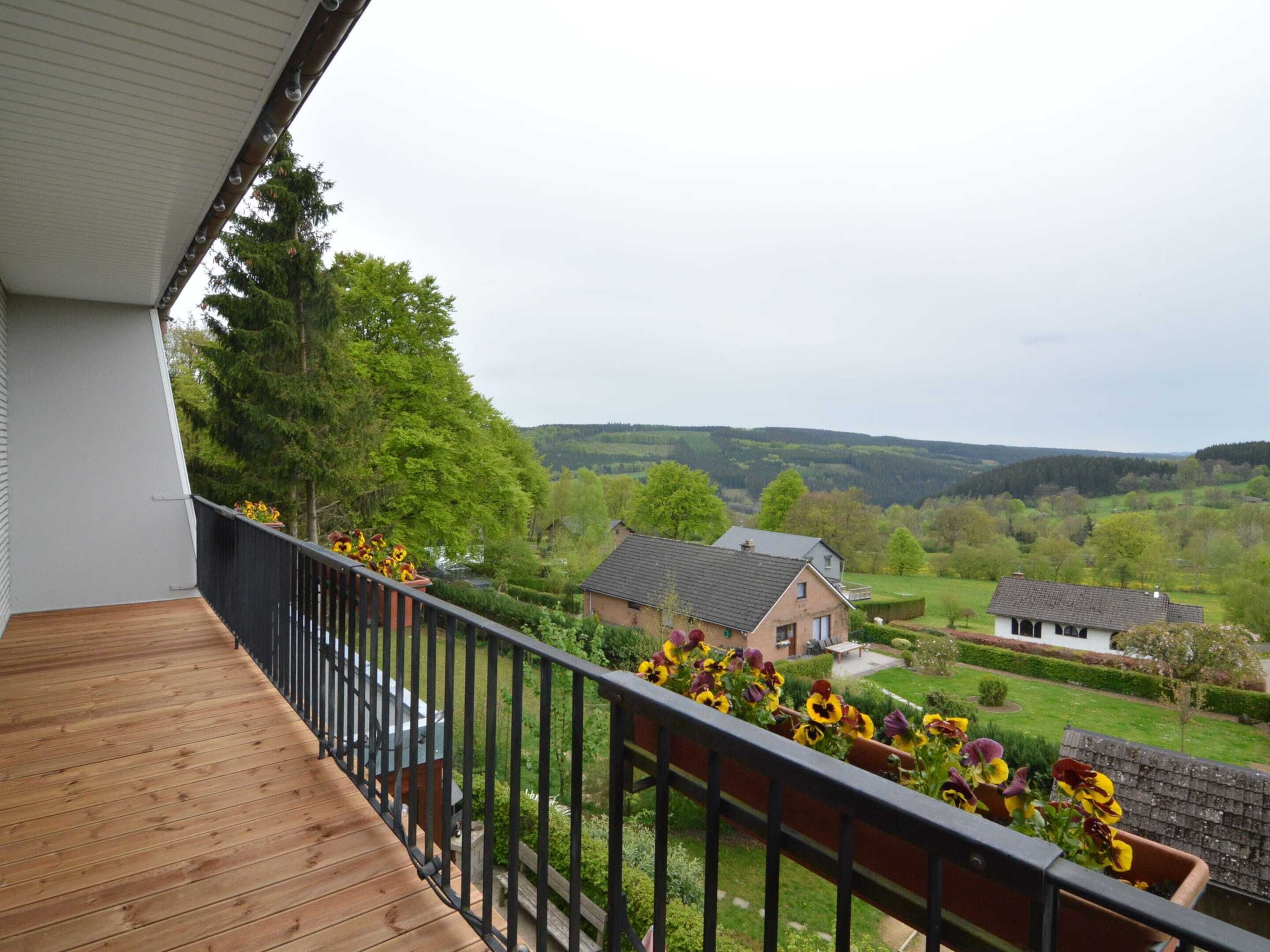 Un balcon en bois, doté d’une rambarde noire, surplombe un paysage rural parsemé de maisons et de verdure.