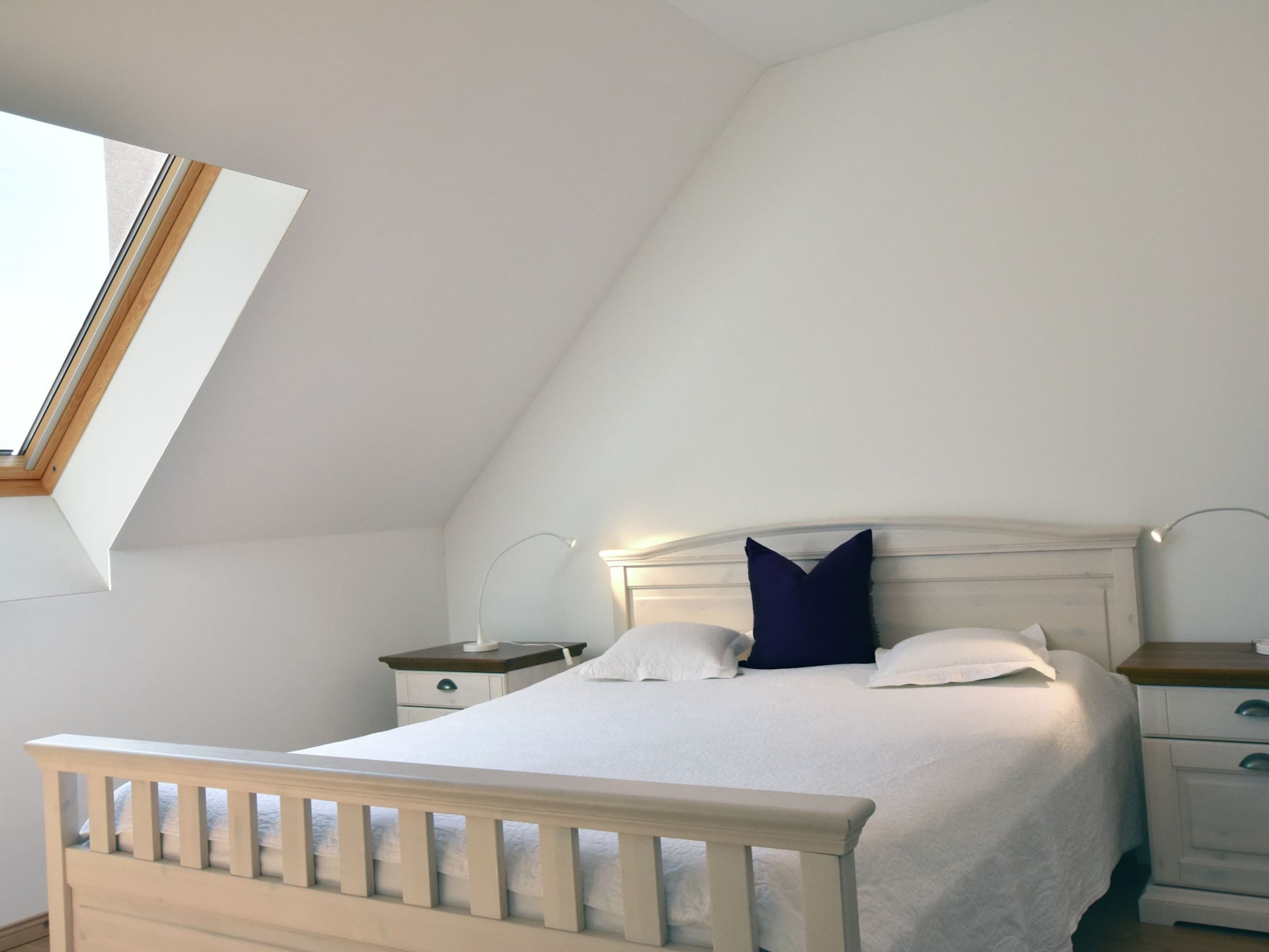 Une chambre avec un plafond en pente, un lit avec une literie blanche, deux tables de chevet et une fenêtre avec un cadre en bois.