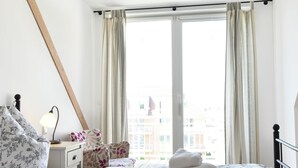 Ein Hotelzimmer mit Bett, Nachttisch mit Lampe, Stuhl und Blick auf Gebäude durch das Fenster.