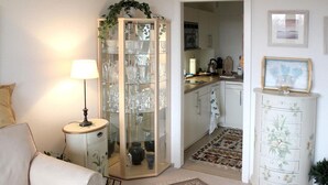A living room with a sofa, a lamp, a glass cabinet, a kitchen area, and a white cabinet with drawers.
