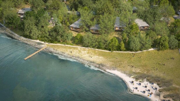 An aerial view of a coastal property with multiple cabins surrounded by dense forest, a sandy beach, and a pier extending into the water.