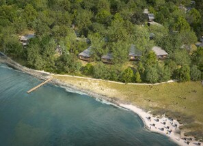 An aerial view of a coastal property with multiple cabins surrounded by dense forest, a sandy beach, and a pier extending into the water. - Sea View Apartment in Glücksburg with Balcony near Sea Beach (Gluecksburg)