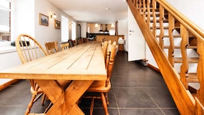 Une longue table à manger en bois, entourée de chaises, un escalier avec des rampes en bois et, au fond, un coin cuisine.