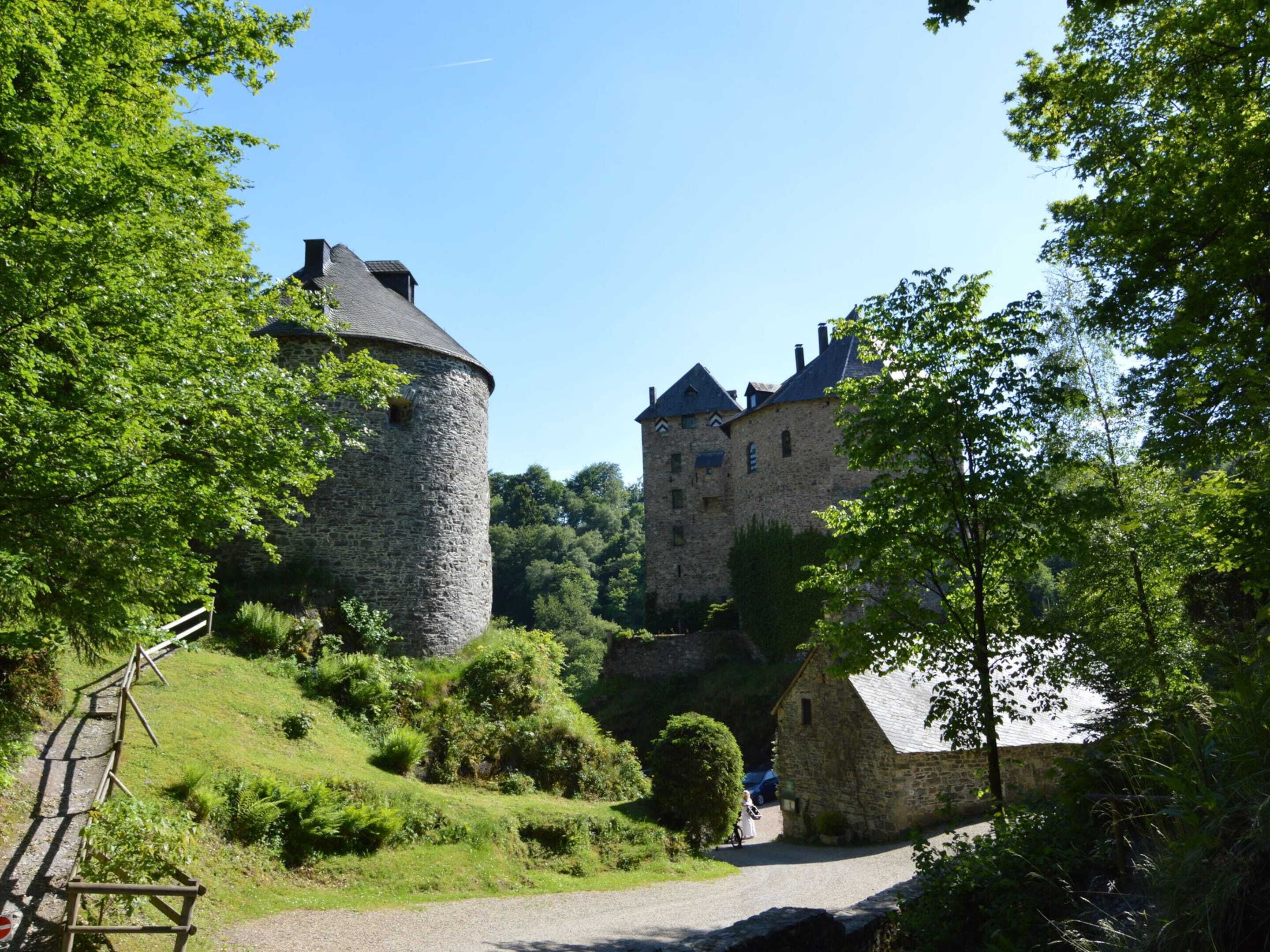 A historic stone castle with a round tower and a larger building with steeply pitched roofs, surrounded by lush greenery and a gravel path.