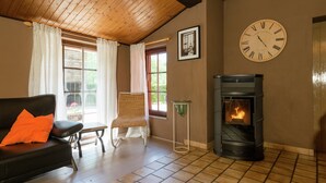 Un salon chaleureux avec une cheminée, un canapé agrémenté d’un coussin orange, un fauteuil en osier et une grande horloge murale.