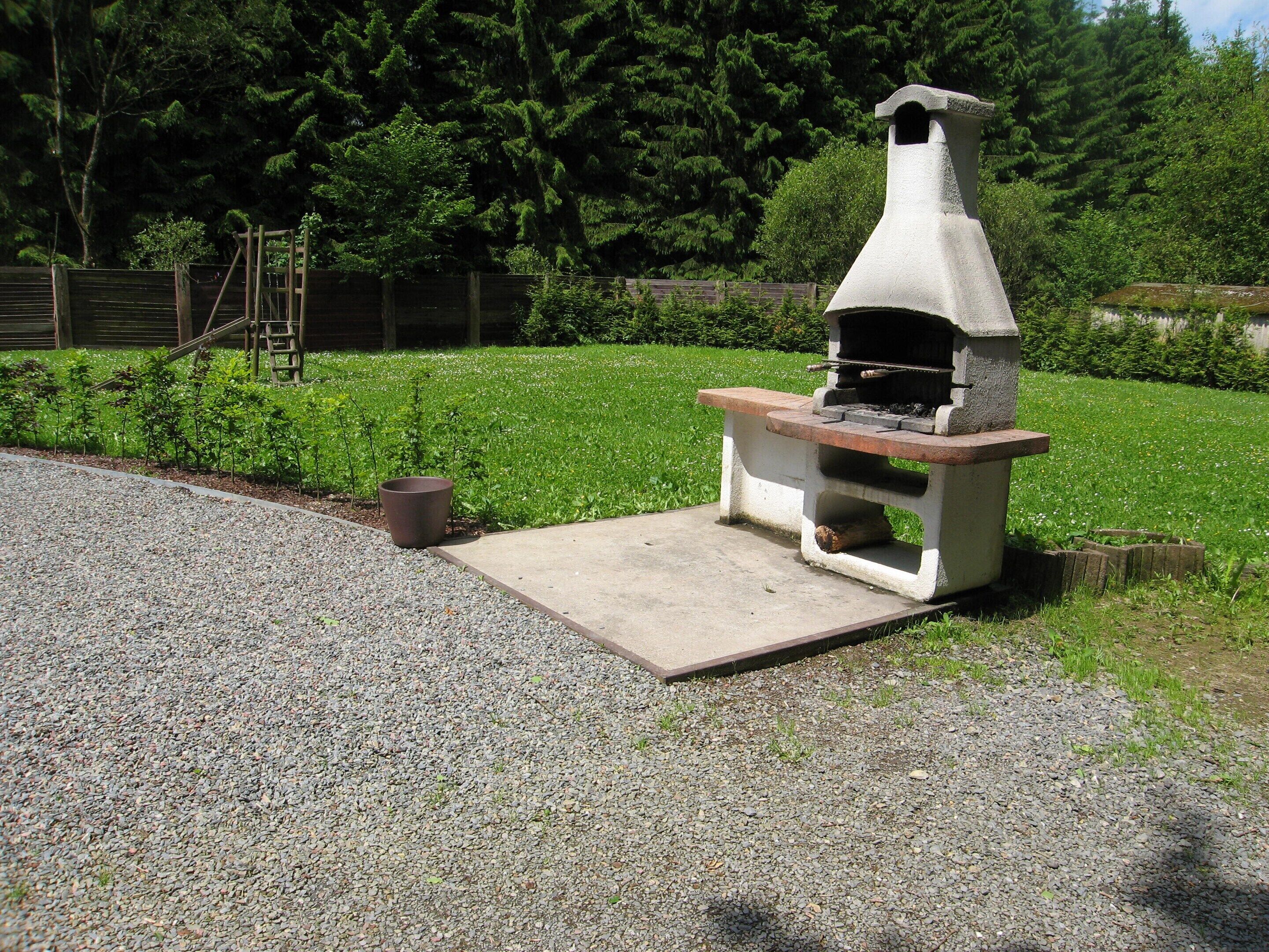 Een betonnen buitenbarbecue met schoorsteen, op een grindpad, met een gazon en bomen op de achtergrond.