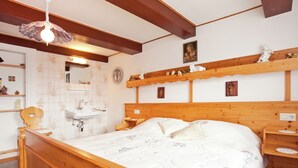 A bedroom with a wooden bed, a wooden headboard, a bedside table, a nightstand, a wall-mounted shelf with decorative items, a mirror, and a hanging light fixture.