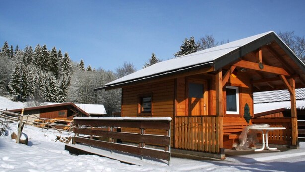 A wooden cabin with a covered porch and a small table, surrounded by snow-covered ground and trees.